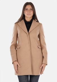 Cappotto di lana beige con un design sartoriale, colletto a revers, bottoni frontali e tasche a pattina. Presenta una silhouette strutturata e una texture liscia.