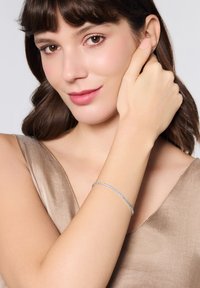 Srebrna bransoletka łańcuszkowa noszona na nadgarstku, z pokręconym wzorem i gładką fakturą, na tle miękkiego, metalicznego materiału.