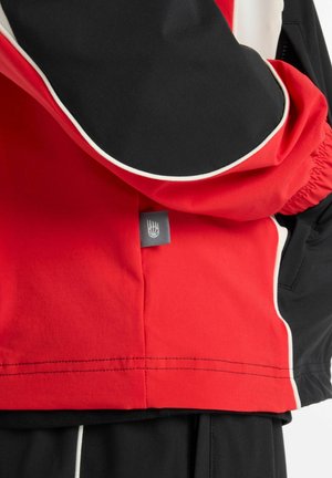 Veste rouge et noire avec des accents blancs, présentant une fermeture éclair, des poignets élastiques et une étiquette de logo près de l'ourlet. Texture lisse, design sportif.