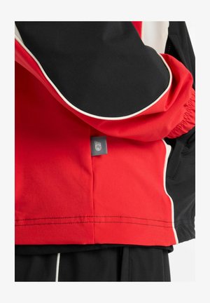 Chaqueta roja y negra con acentos blancos, con cremallera, puños elásticos y una etiqueta de logo cerca del dobladillo. Textura suave, diseño deportivo.