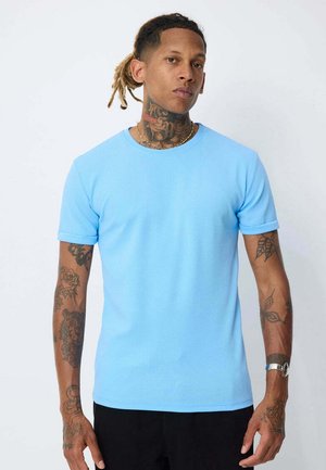 Homme avec dreadlocks et tatouages sur le cou et les bras, portant une chemise bleu clair à manches courtes et une montre-bracelet en argent, se tenant devant un fond uni.