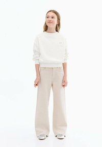 Bílá mikina se žebrovanými manžetami a malým hvězdným logem, spárovaná s béžovými kalhotami širokého střihu a stříbrnými teniskami. Jednoduchý a ležérní design.