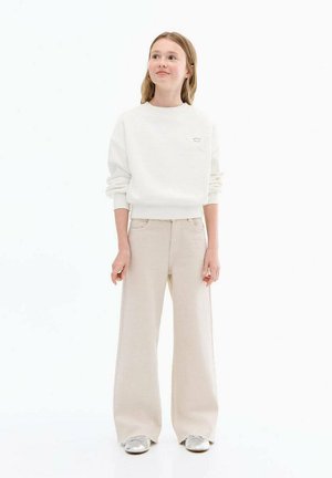 Felpa bianca con polsini a costine e un piccolo logo a stella, abbinata a pantaloni beige a gamba larga e sneaker argentate. Design semplice e casual.