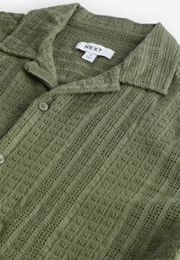 Camisa verde oliva abotonada, confeccionada con un tejido texturizado que presenta un diseño de punto estampado, cuello corto y dos botones en la parte delantera.