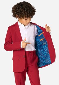 Completo rosso con pantaloni abbinati e giacca, caratterizzato da una fodera a motivo blu. La camicia è bianca e ci sono dettagli in bottone sulla giacca.