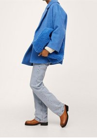 Abrigo de lana azul con un cuello de chal texturizado y esponjoso, llevado sobre un suéter color crema. Jeans rectos de un azul claro y botas de tobillo marrones.
