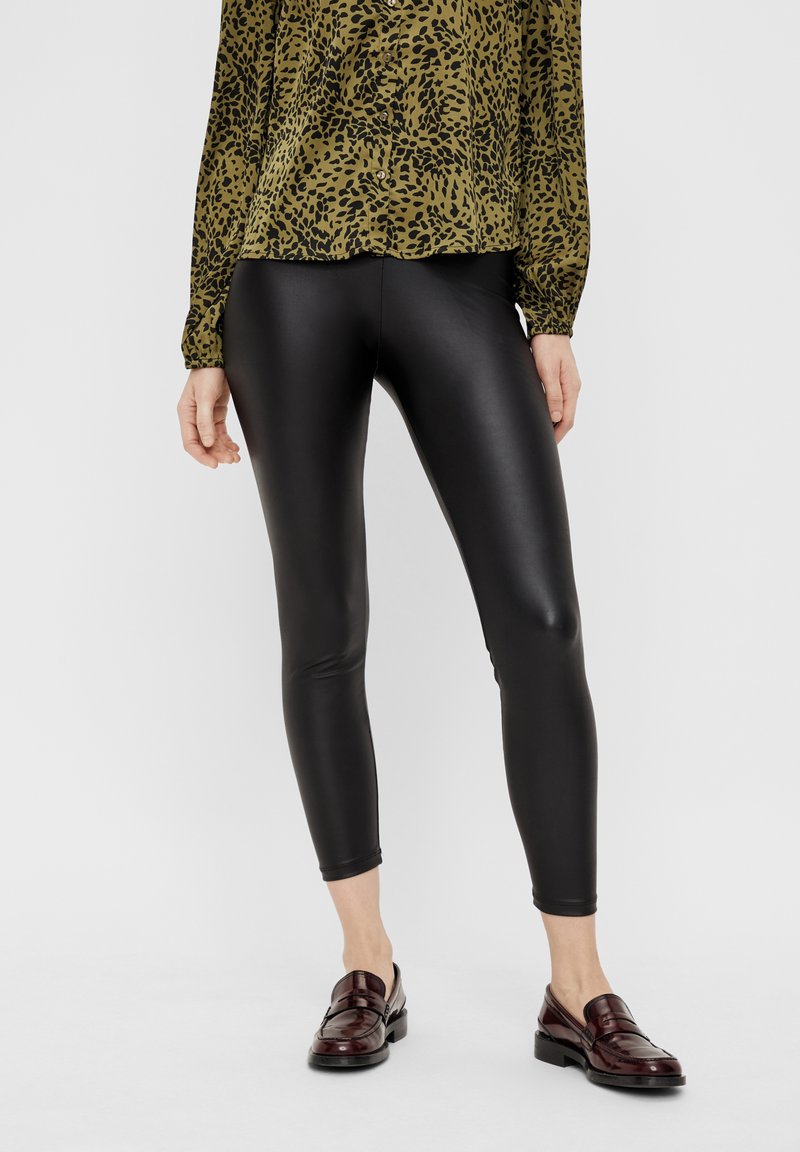 Schwarze, hochglänzende Leggings aus elastischem Material, kombiniert mit einer Bluse im grün-schwarzen Leopardenmuster und kastanienbraunen Lederloafers.