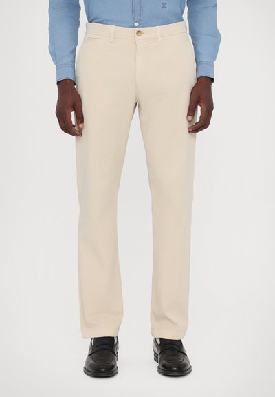 Beige bomuldsbukser med lige snit, der har forlommer og knaplukning, båret med sorte dress sko.