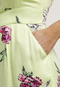 Main reposant dans la poche d'une robe jaune clair avec un imprimé floral rose et vert, montrant la texture du tissu et les détails des coutures.
