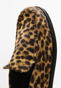 Mocassins en imprimé léopard avec un dessus en fausse fourrure texturée, bout arrondi et semelle en caoutchouc noire. Couture visible le long des bords.
