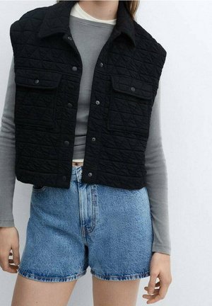 Personne portant une veste matelassée noire par-dessus un haut gris à manches longues et un short en jean bleu sur un fond clair uni.