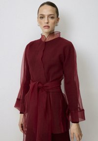 Vestido burdeos con mangas largas de tela transparente, cuello alto con detalle de volantes y cinturón a juego atado en la cintura. Textura de tela suave.