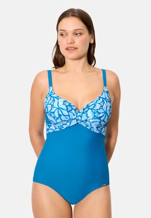 Mujer vistiendo un traje de baño de una pieza azul con la parte superior de patrón floral blanco y azul y tirantes ajustables, de pie frente a un fondo blanco.