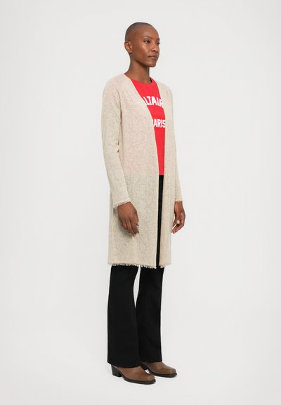 Beige knitted cardigan with an open front, featuring a textured pattern and fringed hem, layered over a red graphic t-shirt and black pants.
