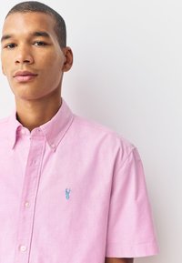 Camicia a maniche corte con bottoni in tessuto rosa chiaro, con logo ricamato di un'aragosta blu sul petto sinistro e collo classico.
