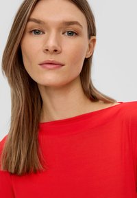 Rotes, weiches Oberteil aus Stoff mit weitem Kragen. Das schulterlange, glatte Haar des Models ergänzt die lebendige Farbe. Neutraler Hintergrund.