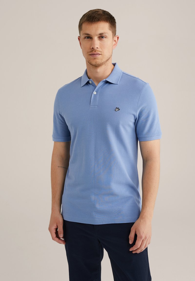 Camisa polo azul claro de tejido suave, con una tapeta de tres botones y un pequeño logo negro bordado en el lado izquierdo del pecho. Mangas cortas.