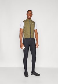 Cyklistický outfit pozostávajúci z olivovo zelenej bezrukávovej vesty so zipsom a logom, skombinovanej s čiernymi priliehavými nohavicami a zodpovedajúcimi cyklistickými topánkami.