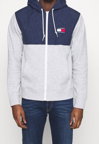 Sudadera con cremallera gris, con un panel superior azul marino, cremallera blanca y detalles de logo. Cuenta con capucha y bolsillos frontales, fabricada con tela suave.