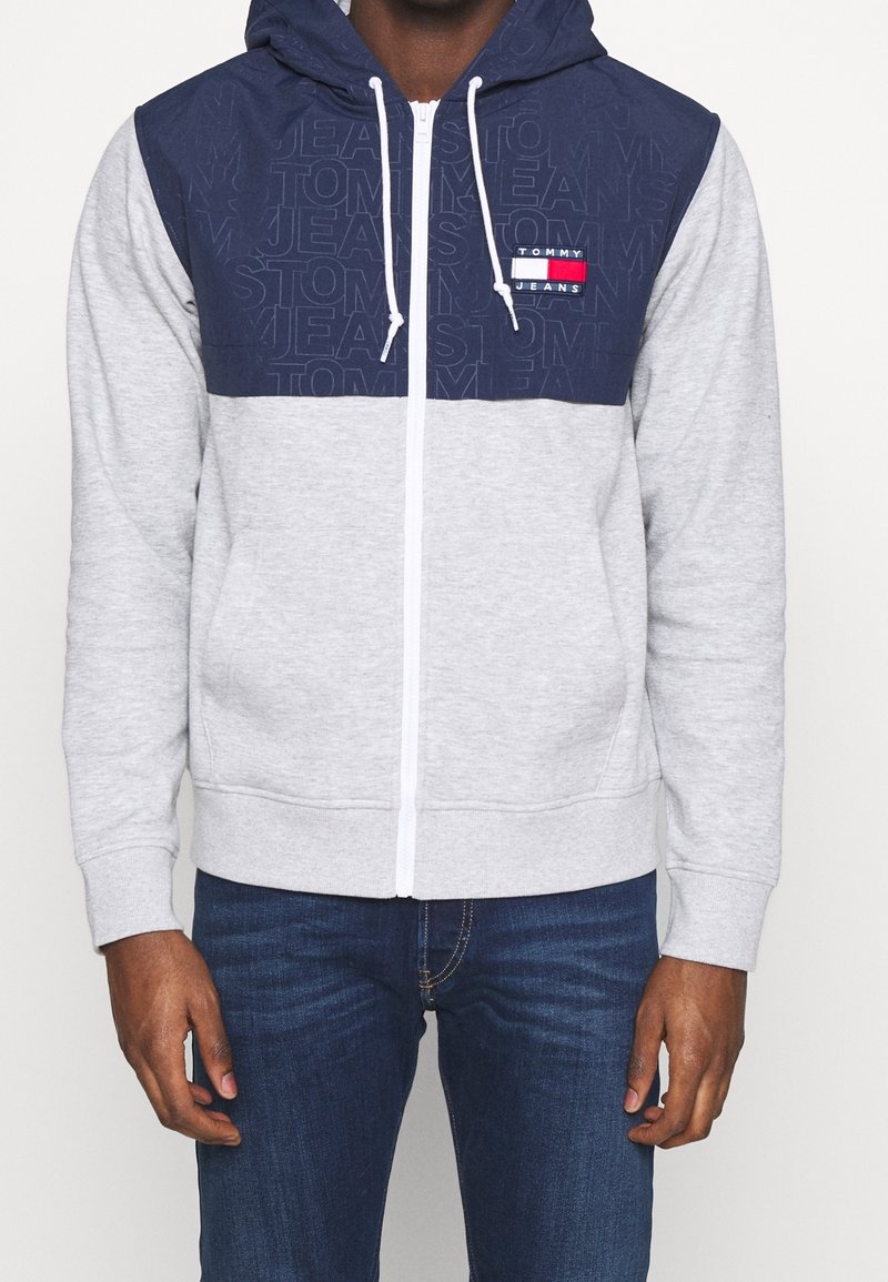 Sudadera con cremallera gris, con un panel superior azul marino, cremallera blanca y detalles de logo. Cuenta con capucha y bolsillos frontales, fabricada con tela suave.