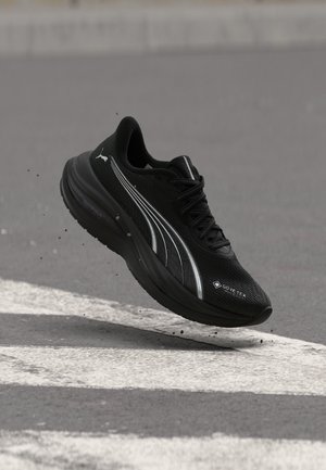 Μαύρο αθλητικό παπούτσι Puma με λευκές ρίγες και ετικέτα Gore-Tex, αιωρείται πάνω από δρόμο με λευκές διαβάσεις πεζών.