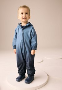 Mėlynas ir tamsiai mėlynas neperšlampamas kūdikių kombinezonas su priekinio užtrauktuku, elastingomis rankogaliais ir derančiais mėlynais guminiais batais ant neutralaus fono.