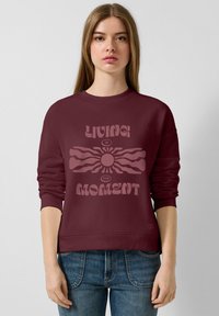 Sweatshirt bordeaux à col rond et manches longues, avec un design graphique central comprenant le texte "VIVRE L'INSTANT".