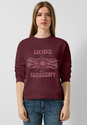 Sweatshirt bordeaux à col rond et manches longues, avec un design graphique central comprenant le texte "VIVRE L'INSTANT".