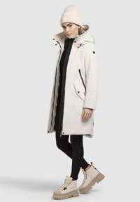 Witte capuchonjas met ritszakken, zwarte accenten, gecombineerd met een zwarte jurk, zwarte leggings en beige laarzen met dikke zolen. Gehaakte beige beanie.