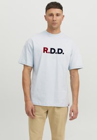 Vaaleansininen puuvillapaita, jossa on rohkea "R.D.D." -teksti punaisena ja tummansinisenä. Lyhyet hihat ja rento malli, yhdistettynä beigeihin housuihin.