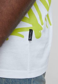 White cotton t-shirt featuring a large, bright green graphic design. Includes a small black label on the side seam.