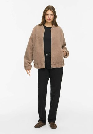 Frau steht und trägt eine braune Fleecejacke mit Reißverschluss, ein schwarzes Shirt und eine schwarze Hose sowie braune Mokassins vor einem schlichten weißen Hintergrund.