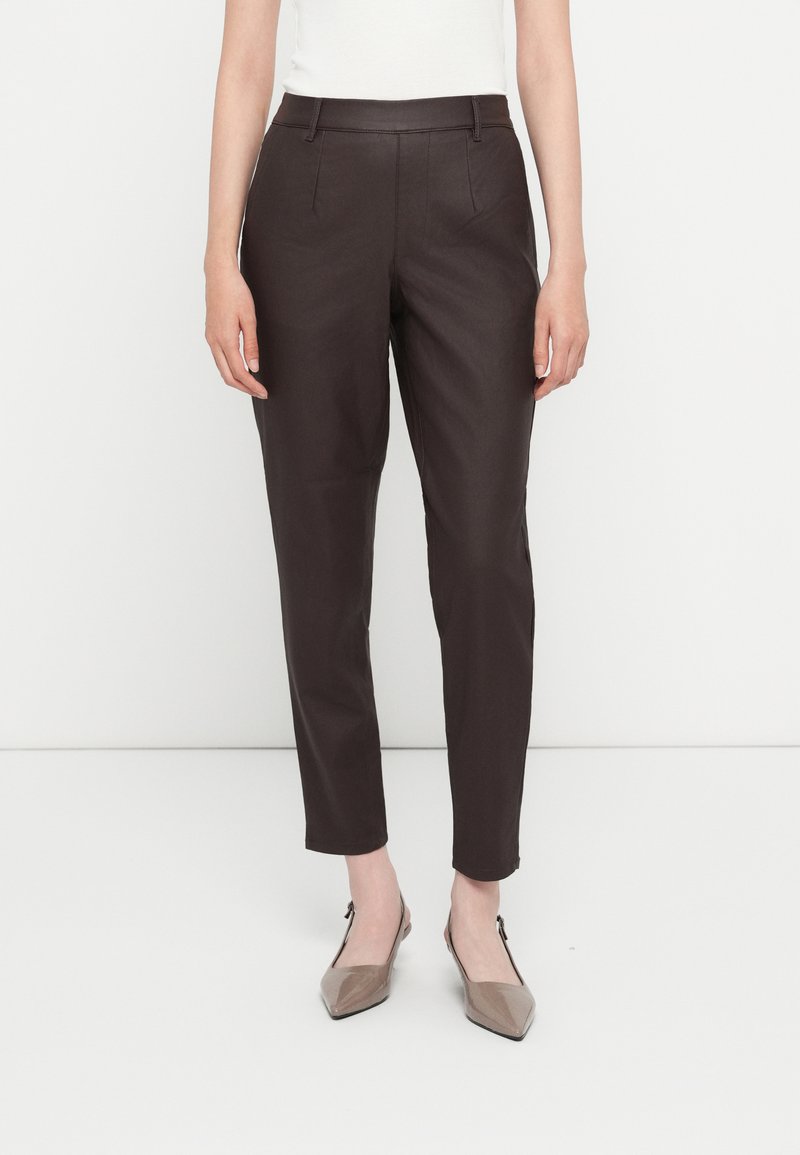 Pantalones marrones ajustados con una textura suave, que presentan pliegues frontales y una cinturilla plana. Combinados con unos zapatos planos beige de punta afilada.