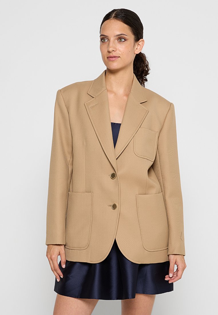 Beige textuur blazer met een notch lapel, twee frontzakken en goudkleurige knopen, gedragen over een marineblauwe jurk.