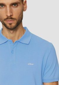 Hellblaues Polo-Shirt aus strukturiertem Stoff, mit Kragen und Knopfleiste mit zwei Knöpfen. Kleines weißes Logo auf der Brust.