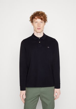 Donkerblauwe lange mouwen polo shirt met een drieknoopsluiting, zachte textuur en een klein geborduurd logo op de borst. Gecombineerd met lichtgroene broek.
