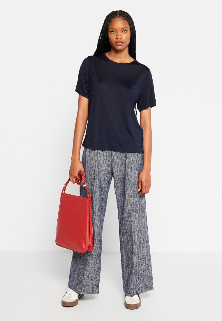 Marineblaues Kurzarm-T-Shirt kombiniert mit weiten gemusterten Hosen, hält eine rote Handtasche. Weiße Sneaker mit braunen Sohlen.