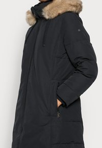 Polstret sort parka med hætte med pelskant, frontlynlås og sidelommer. Har en glat overflade og quiltet struktur.