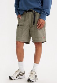 Kaki cargoshorts met twee zijzakken, een riem en een rood merkembleem, gecombineerd met zilverkleurige sportschoenen en witte sokken.