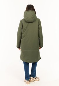 Olijfgroene gewatteerde jas met een capuchon, lang ontwerp, geribde manchetten en zijsplitsen. Draagt over blauwe jeans en beige sneakers.