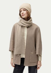 Beige ullponcho med vida ärmar, två framfickor och en guldslogo som accent. Matchas med en passande ribbad mössa och scarf.