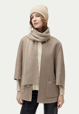 Beige wool poncho with wide sleeves, two front pockets, and a gold logo accent. Paired with a matching ribbed beanie and scarf.