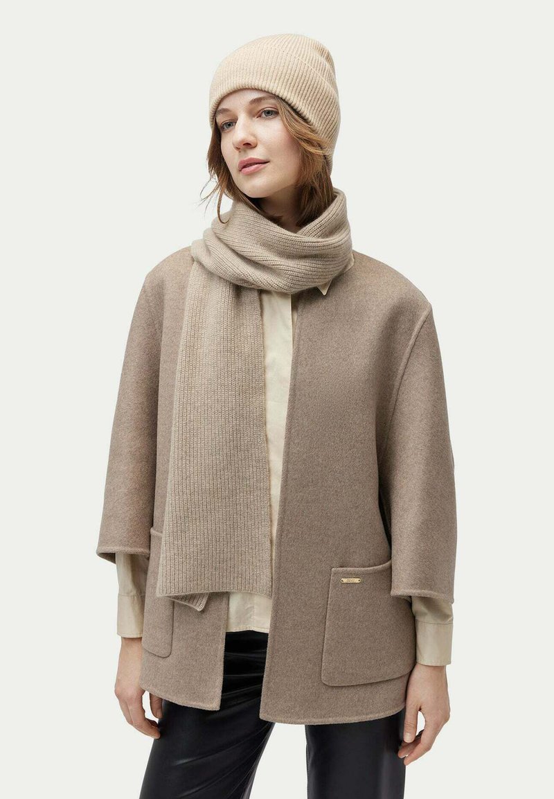 Beige ullponcho med vida ärmar, två framfickor och en guldslogo som accent. Matchas med en passande ribbad mössa och scarf.