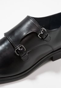 Zwarte leren schoen met een gladde afwerking, voorzien van twee gespen, een ronde neus en een lage profiel. Stevige zool met een lichte textuur.