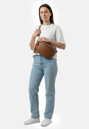 Young woman in white t-shirt and light jeans holds large brown leather crossbody bag with tassel, standing against white background.