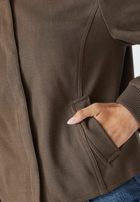 Brązowa, prążkowana kurtka z zbliżeniem na rękę w bocznej kieszeni. Tkanina ma teksturowany wygląd i miękkie wykończenie.