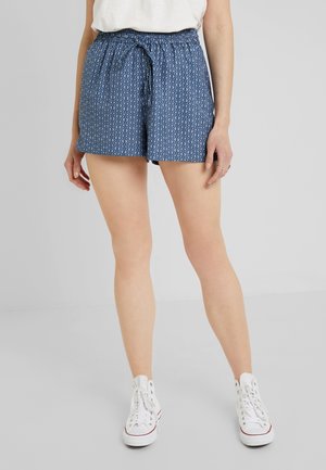 Kvinde iført blå mønstrede shorts med snøring, hvide sneakers og en hvid top, stående foran en ensfarvet grå baggrund.