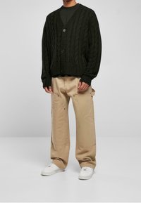 Mörkgrön tjockstickad kofta med V-hals, med knapplist framtill och ribbat avslut, parat med beige vida byxor och vita sneakers.