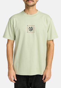 Světlezelené bavlněné tričko s centrálním grafickým designem v černé a béžové barvě. Krátké rukávy a volný střih.