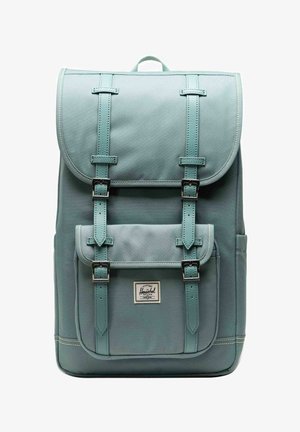 Sac à dos en tissu bleu clair avec poche avant et deux sangles à boucle, poignée de transport sur le dessus, et étiquette de marque Herschel sur la poche.